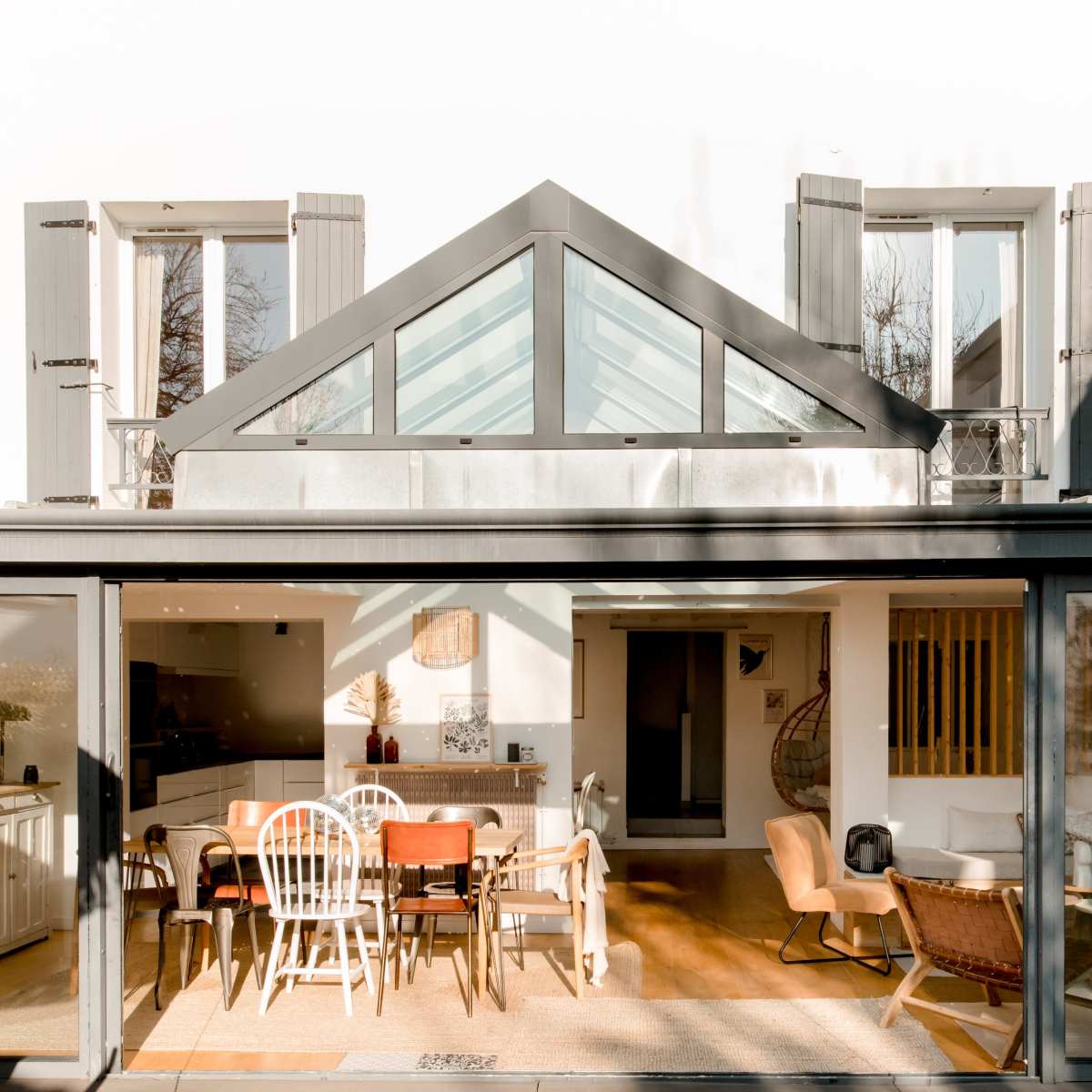 A modern house with large glass doors and a spacious dining area, featuring a wooden table and various chairs, under a sloped glass roof.