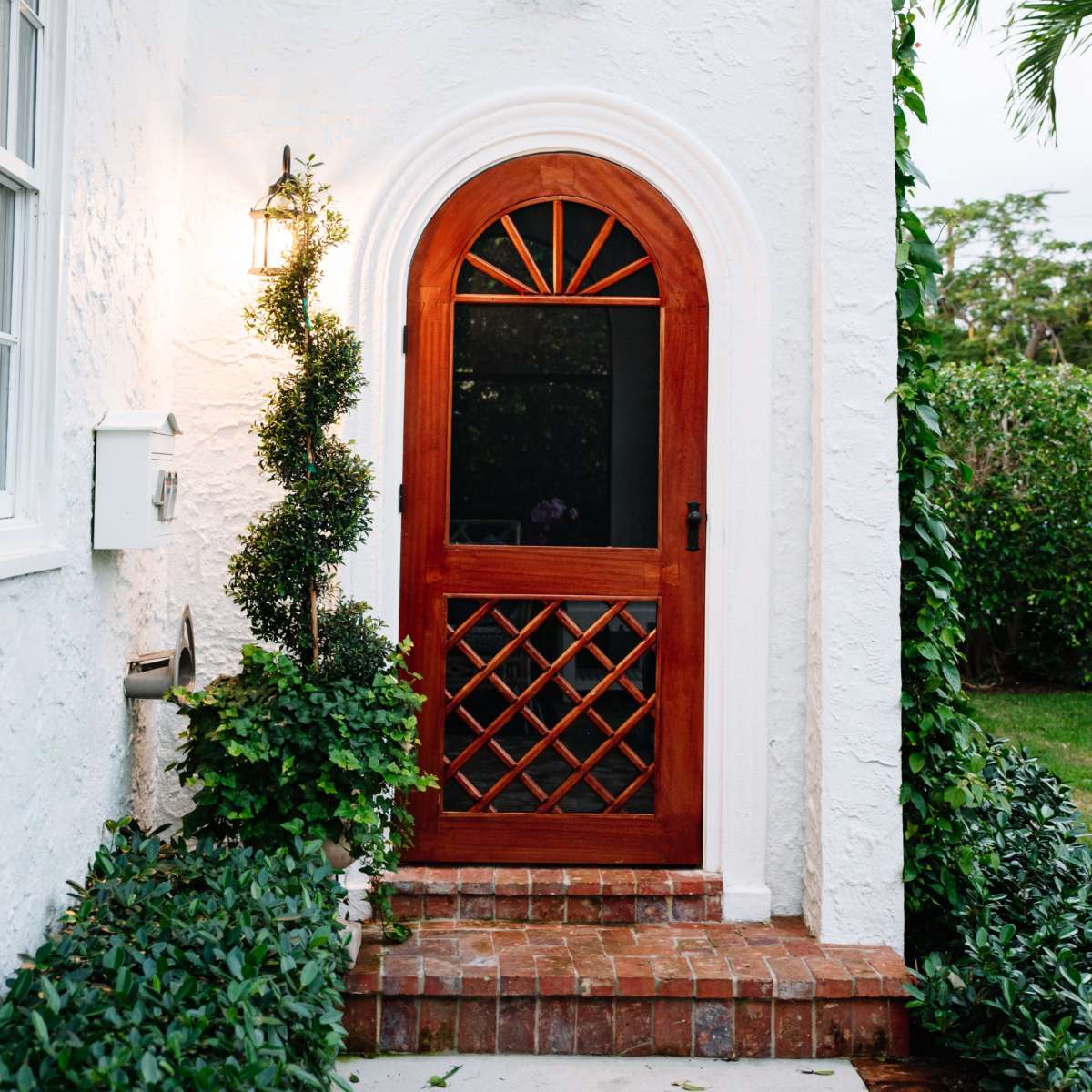 A white stucco house entrance featuring a wooden arched door, flanked by greenery and a wall-mounted lantern.