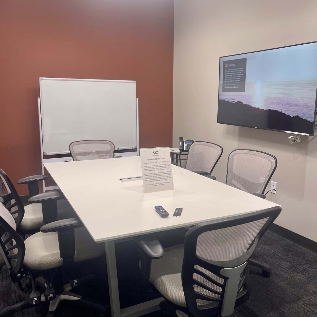 A small meeting room with a rectangular table, six chairs, a whiteboard, and a wall-mounted television.