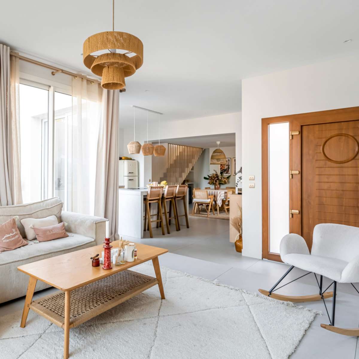 A cozy living room features a light-colored sofa, a wooden coffee table, and a rocking chair, with large windows and a view of an adjoining dining area.