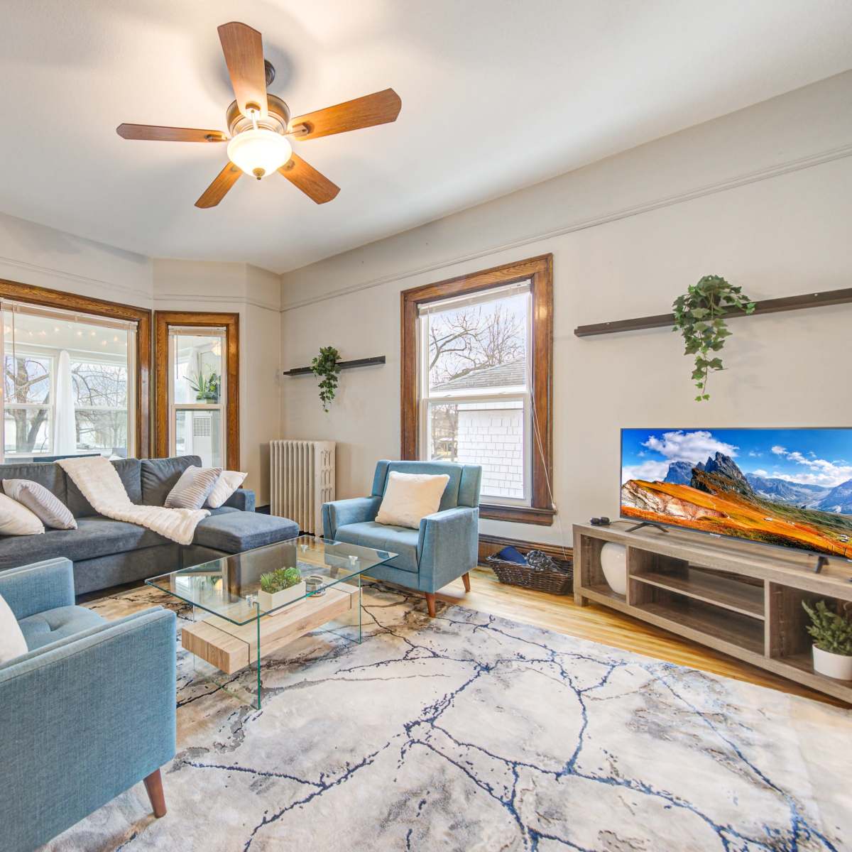 A bright living room features large windows, a ceiling fan, a glass coffee table, and modern furniture with a television displaying a mountain landscape.