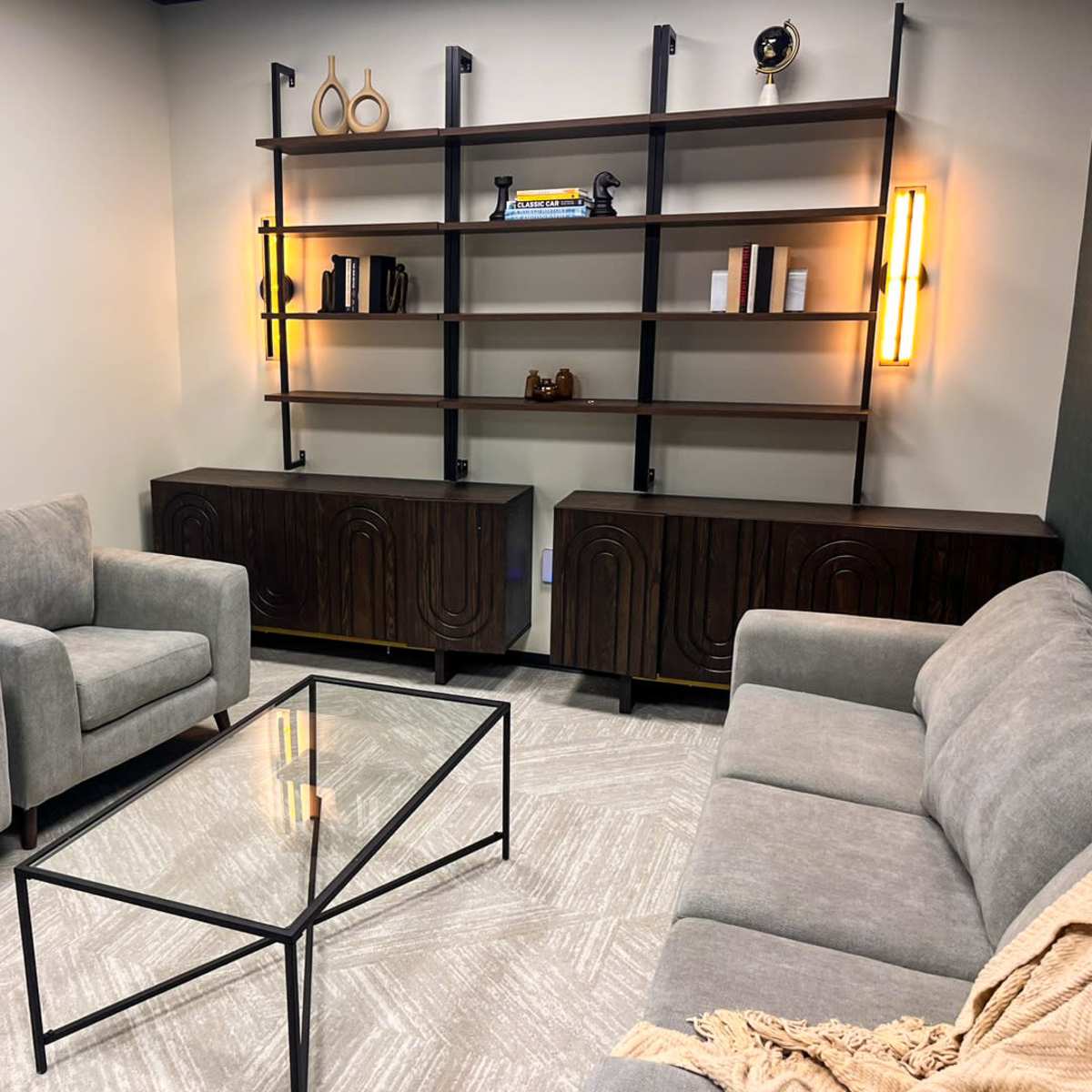A minimalist lounge area with four gray chairs arranged around a glass coffee table, complemented by a dark wooden shelving unit and two illuminated wall fixtures.