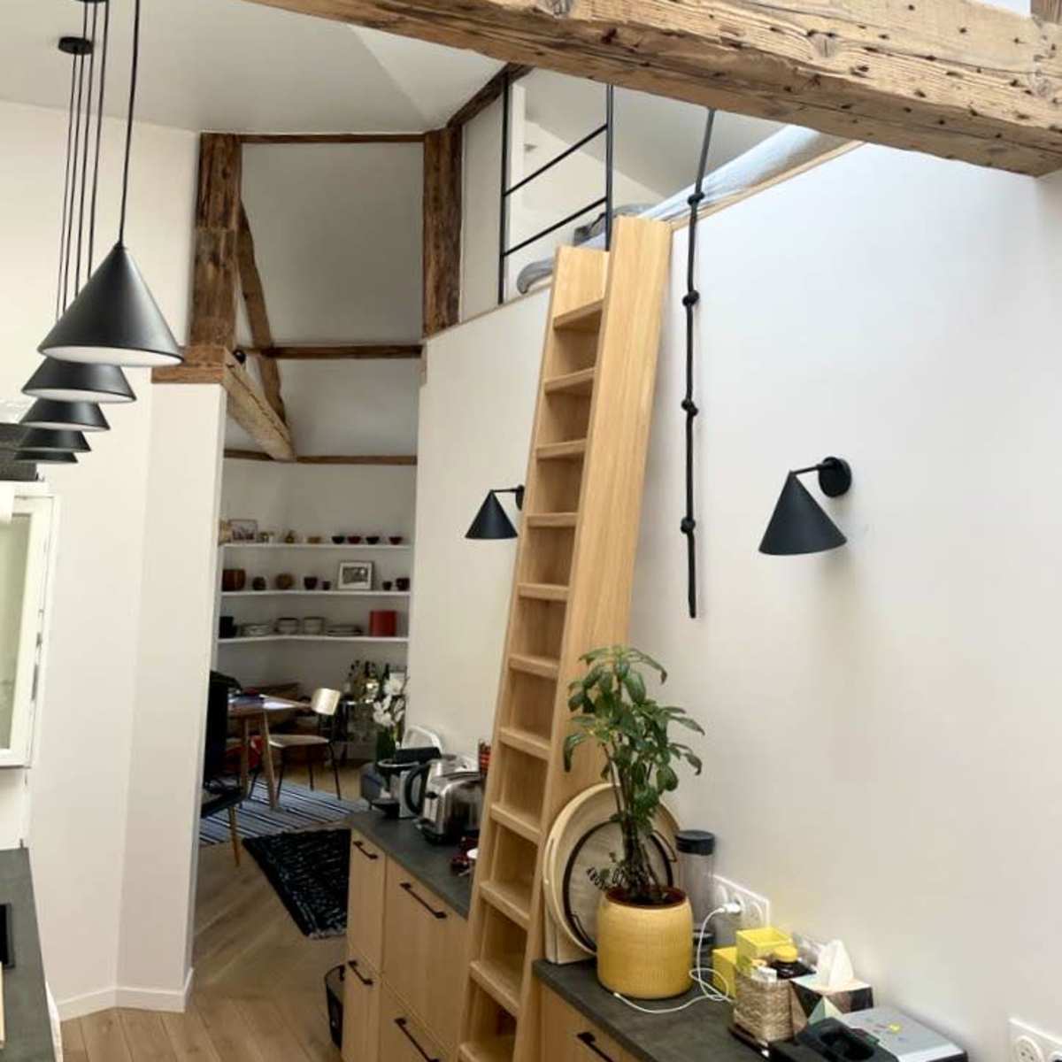 A modern kitchen with wooden beams, a ladder leading to an upper level, and pendant lights hanging above a countertop.