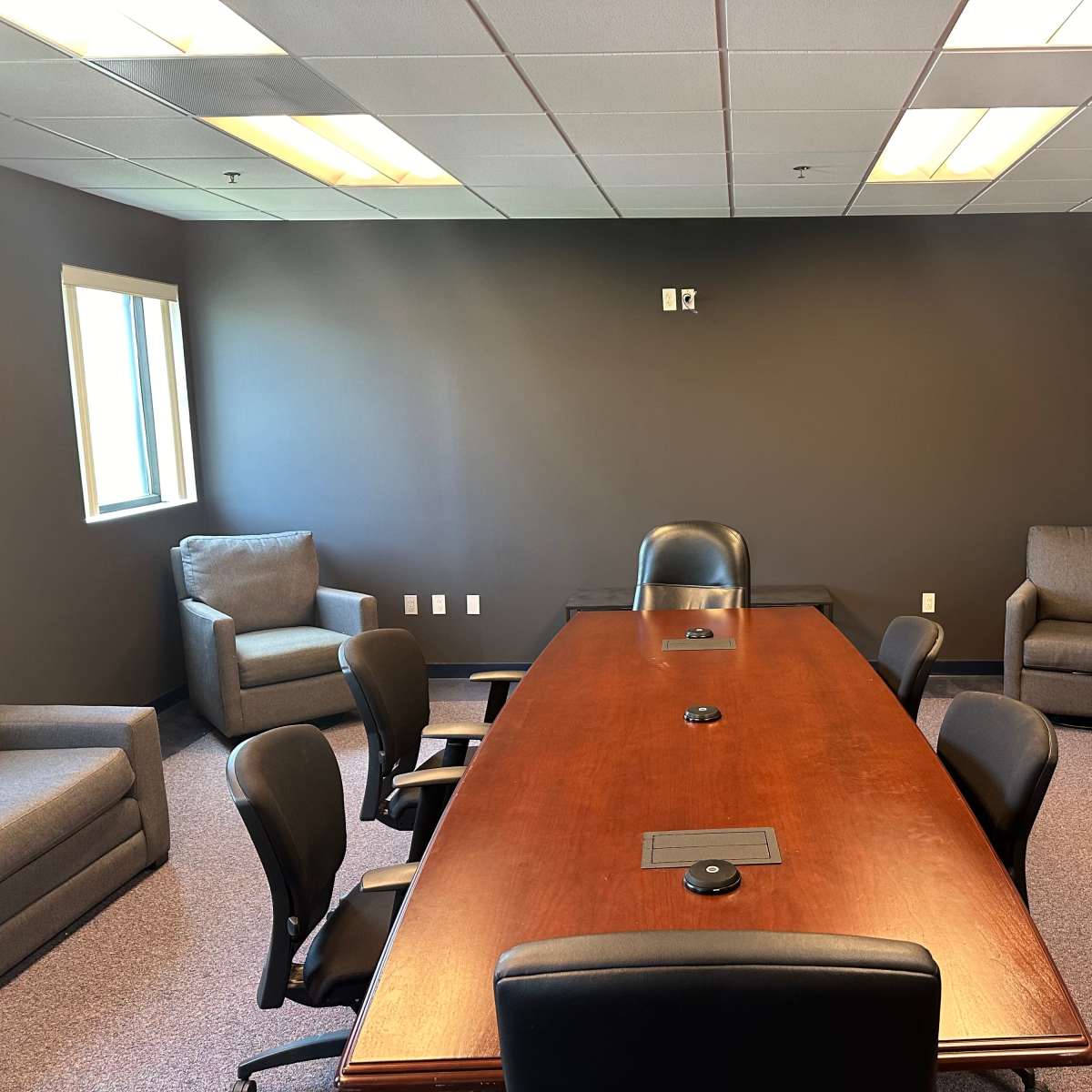 A meeting room with a large wooden table surrounded by black office chairs, flanked by two armchairs and a window.