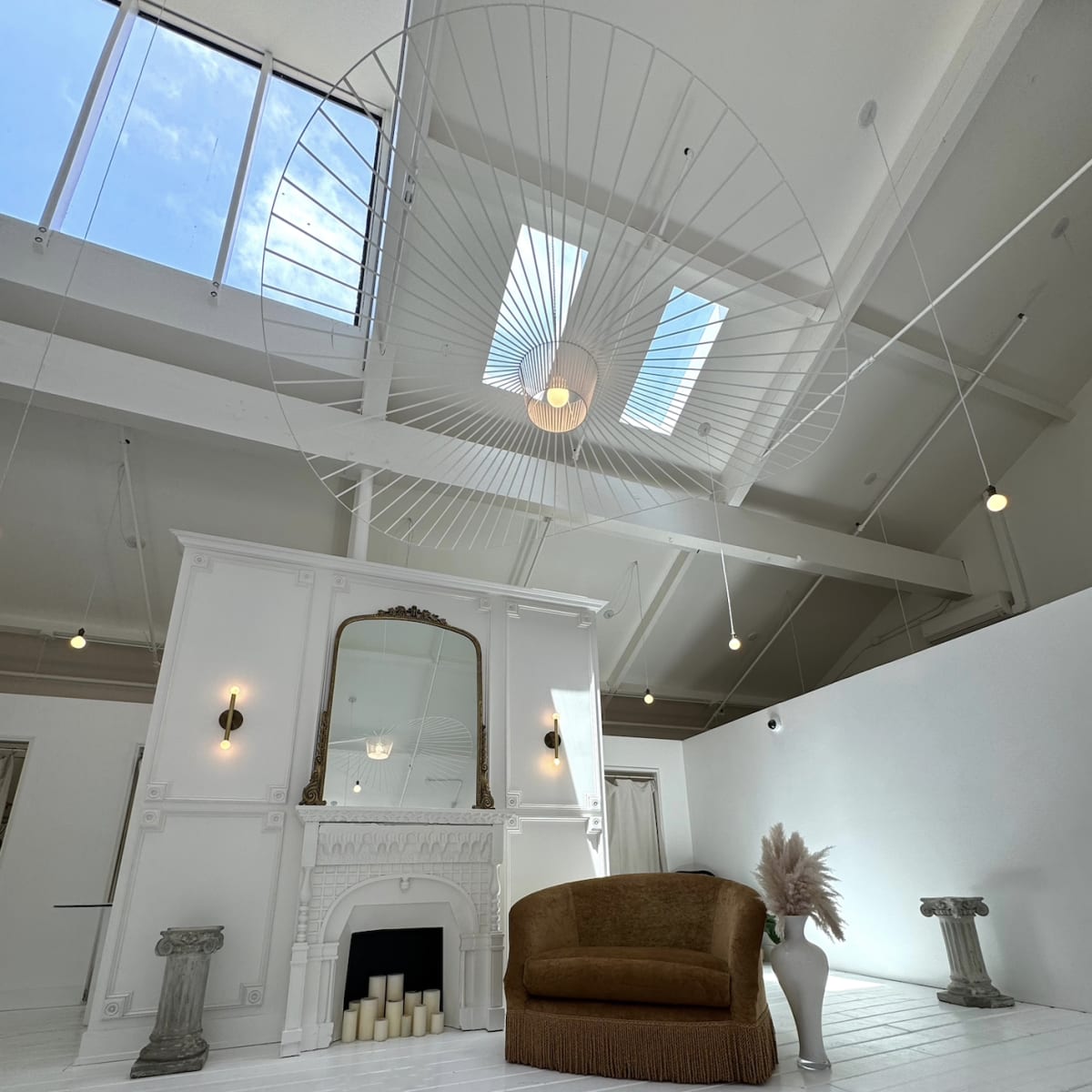 A spacious room with white wooden floors, a large mirror above a decorative fireplace, a brown sofa, and natural light coming through a skylight.