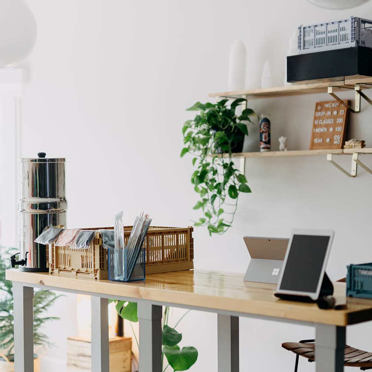 A modern workspace features a long wooden table with a beverage dispenser and organizational baskets, against a backdrop of greenery and minimalist decor.