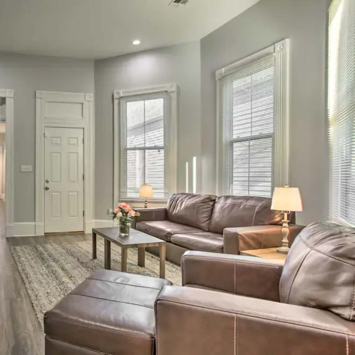 A modern living room featuring two brown leather sofas, a coffee table, a wall-mounted TV, and bright windows, with a hallway visible in the background.
