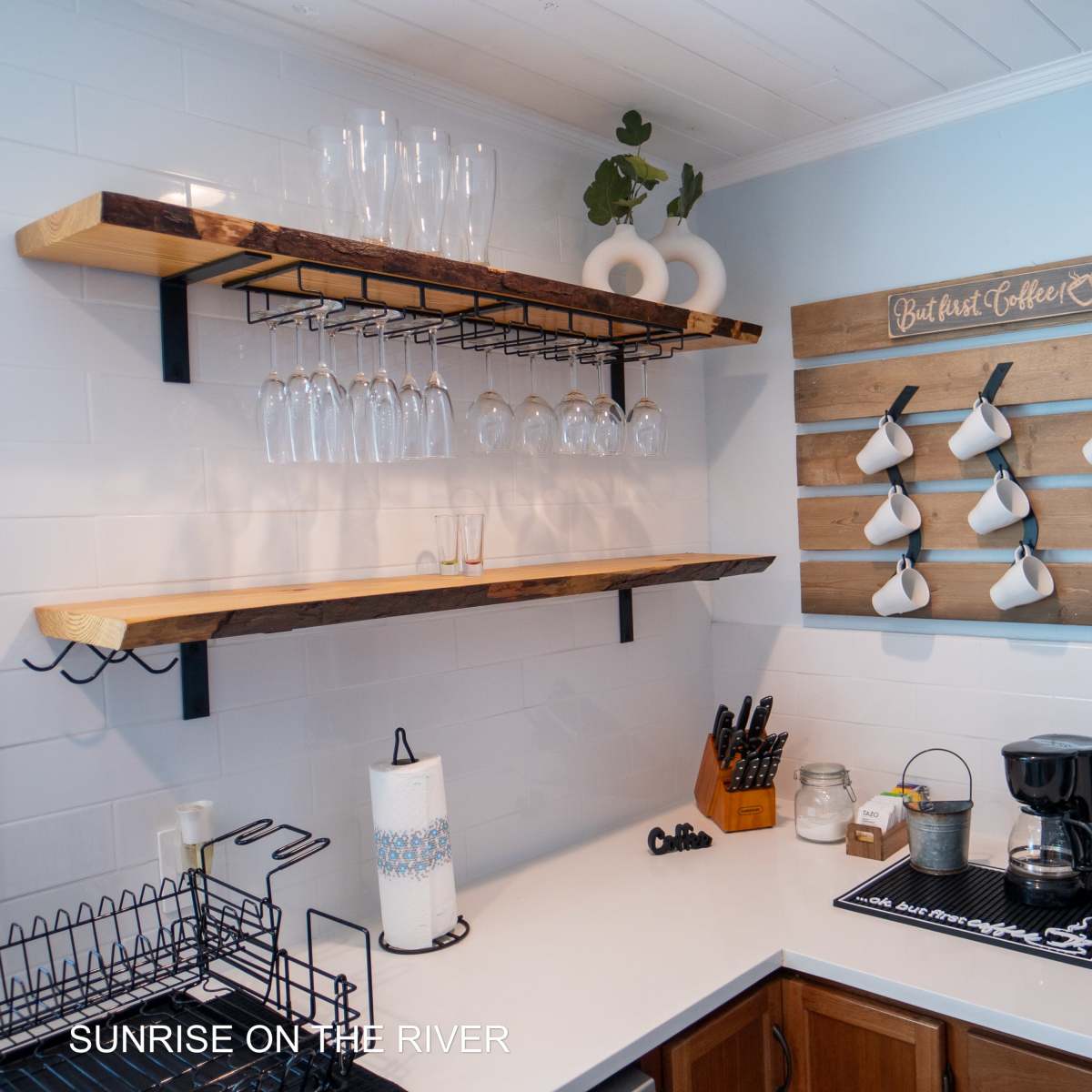 A kitchen corner with two wooden shelves displaying glassware and a wall-mounted rack holding white mugs, alongside a coffee maker and utensils on the counter.