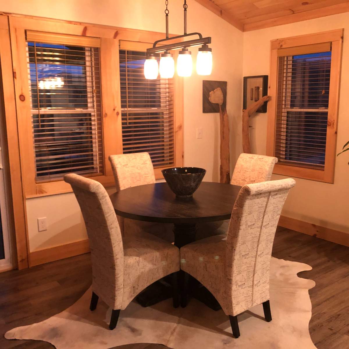 A round dining table with four upholstered chairs is situated in a bright room featuring large windows and wooden accents.