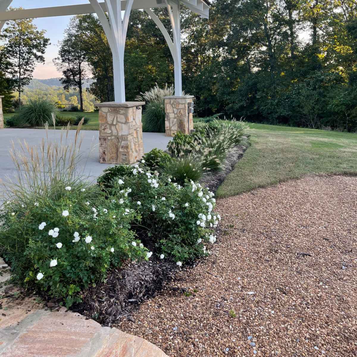 A covered patio area with stone pillars surrounded by landscaped greenery and white flowers, set against a backdrop of trees and a clear sky.