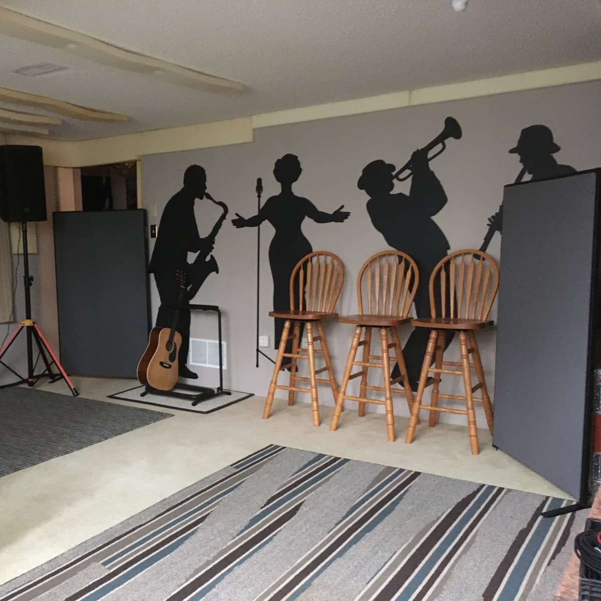 A music practice room featuring silhouettes of musicians on the walls, three wooden stools, an acoustic guitar, and a sound system setup.