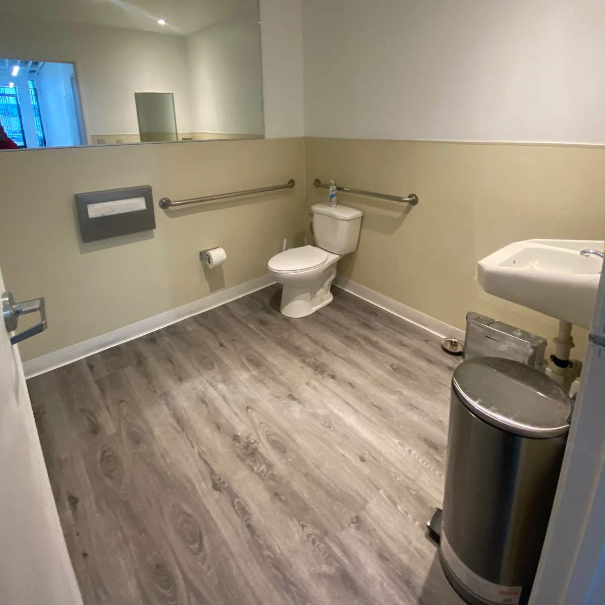 A modern restroom with a toilet, sink, grab bars, a mirror, and a waste bin on a wooden floor.