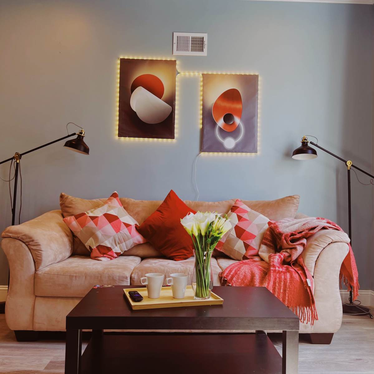 A cozy living room featuring a beige sofa adorned with colorful cushions, a wooden coffee table, and artwork on the wall illuminated by lights.