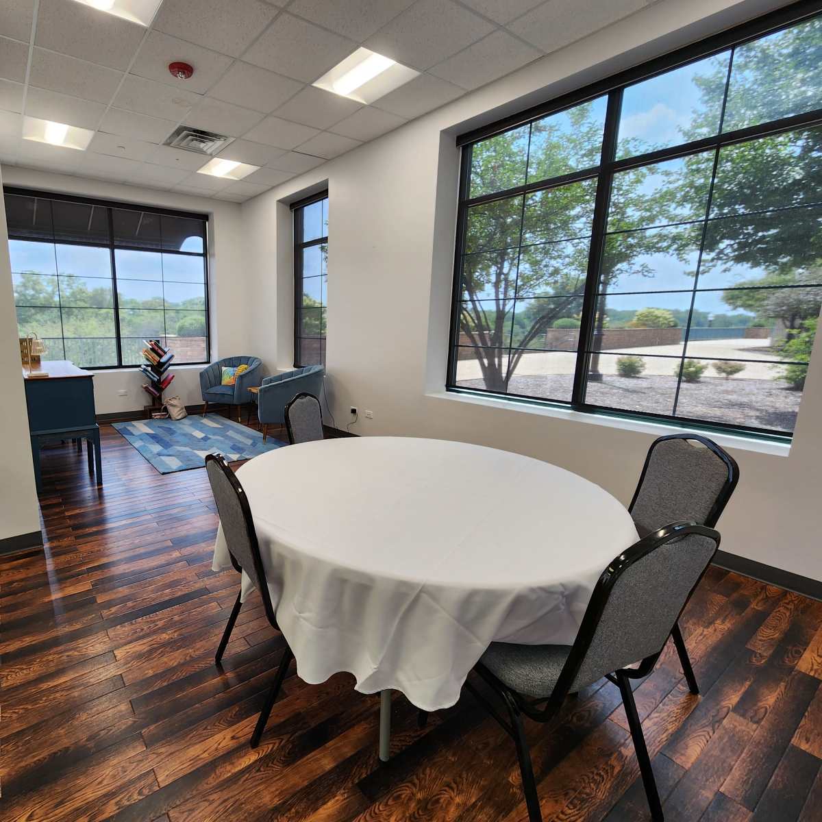 A well-lit meeting room featuring a round table with chairs, large windows overlooking a landscape, and a small seating area.