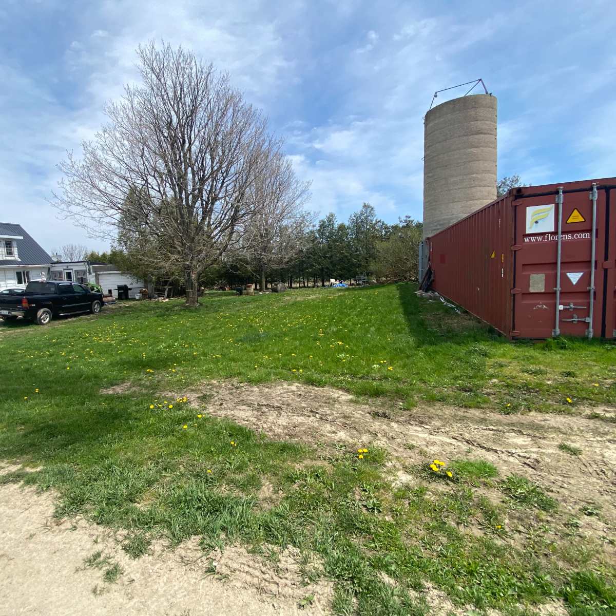 A gravel driveway leads to a house with a dark roof, parked cars, a large tree, and a cylindrical storage silo on a grassy area.