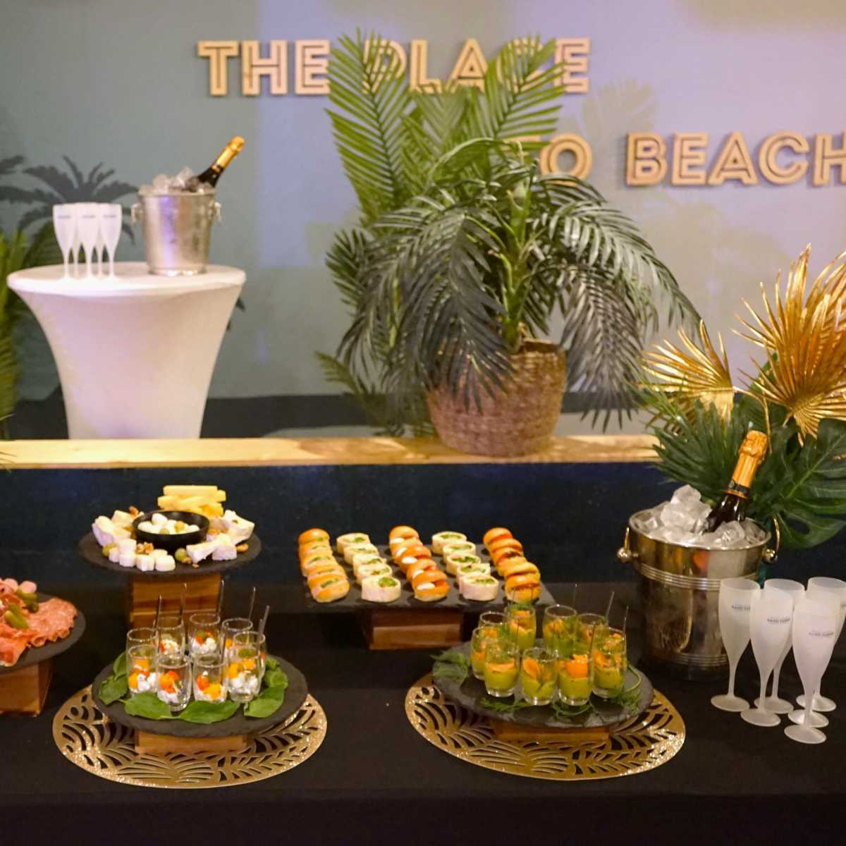 A buffet display features a variety of sushi, seafood, and cocktails set against a backdrop of decorative plants and a champagne bucket.