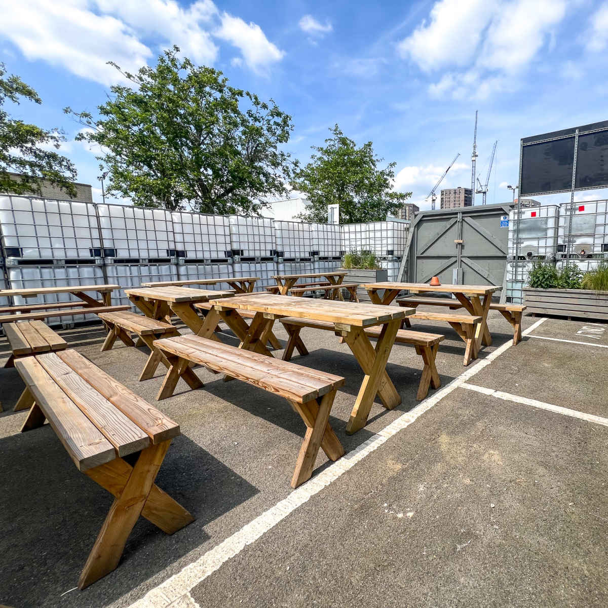 An outdoor seating area with wooden picnic tables surrounded by industrial containers and greenery under a blue sky.