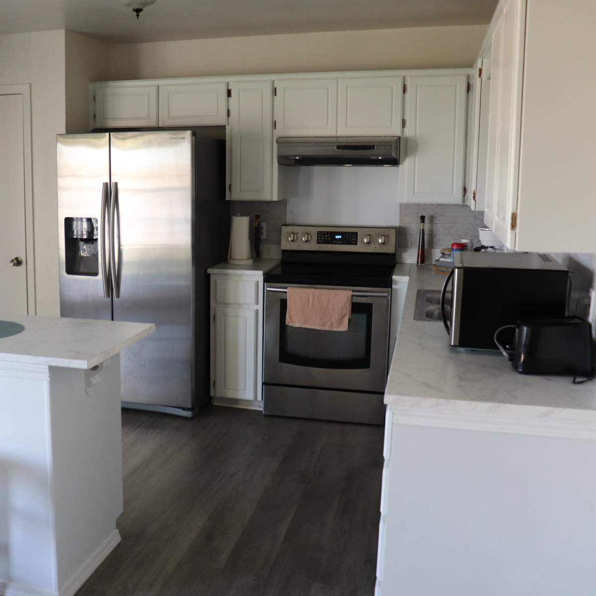 A modern kitchen features stainless steel appliances, white cabinetry, and minimal furnishings including a small table and stools.