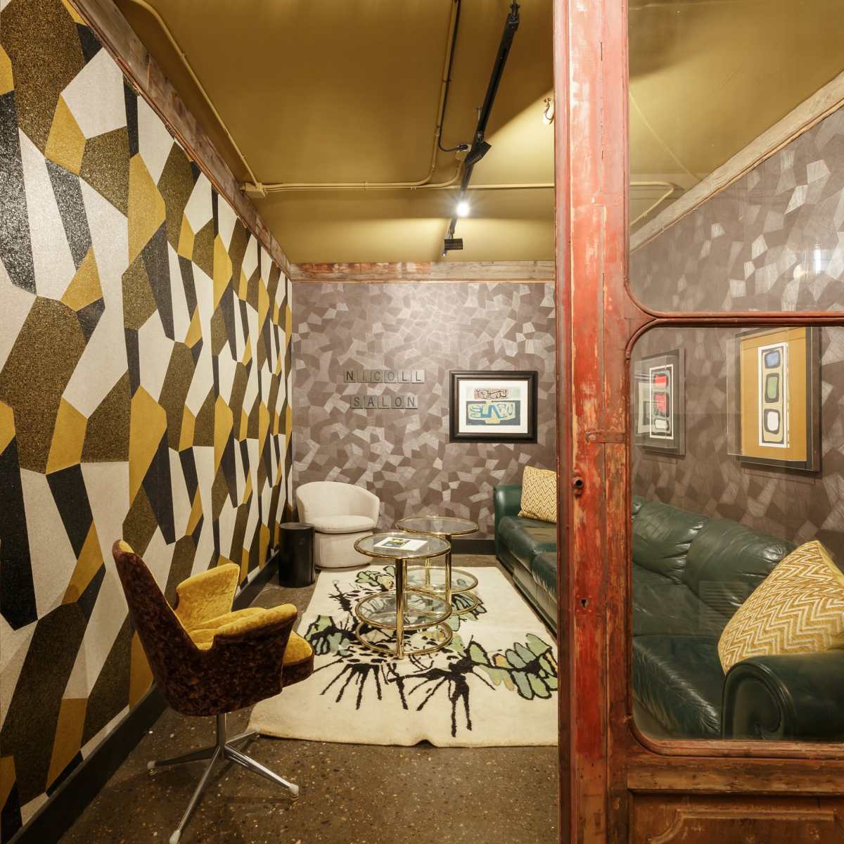 The room features a combination of geometric patterned walls, a green leather sofa, a round coffee table, and a yellow-accented chair, enclosed by a glass door.