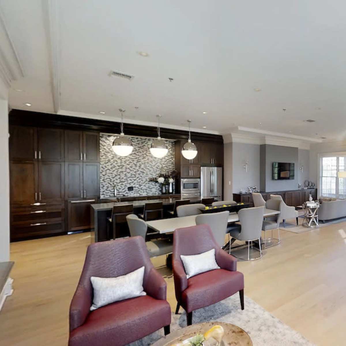 A spacious modern lounge area features a kitchen in the background, seating arrangements, and large windows allowing natural light to fill the room.