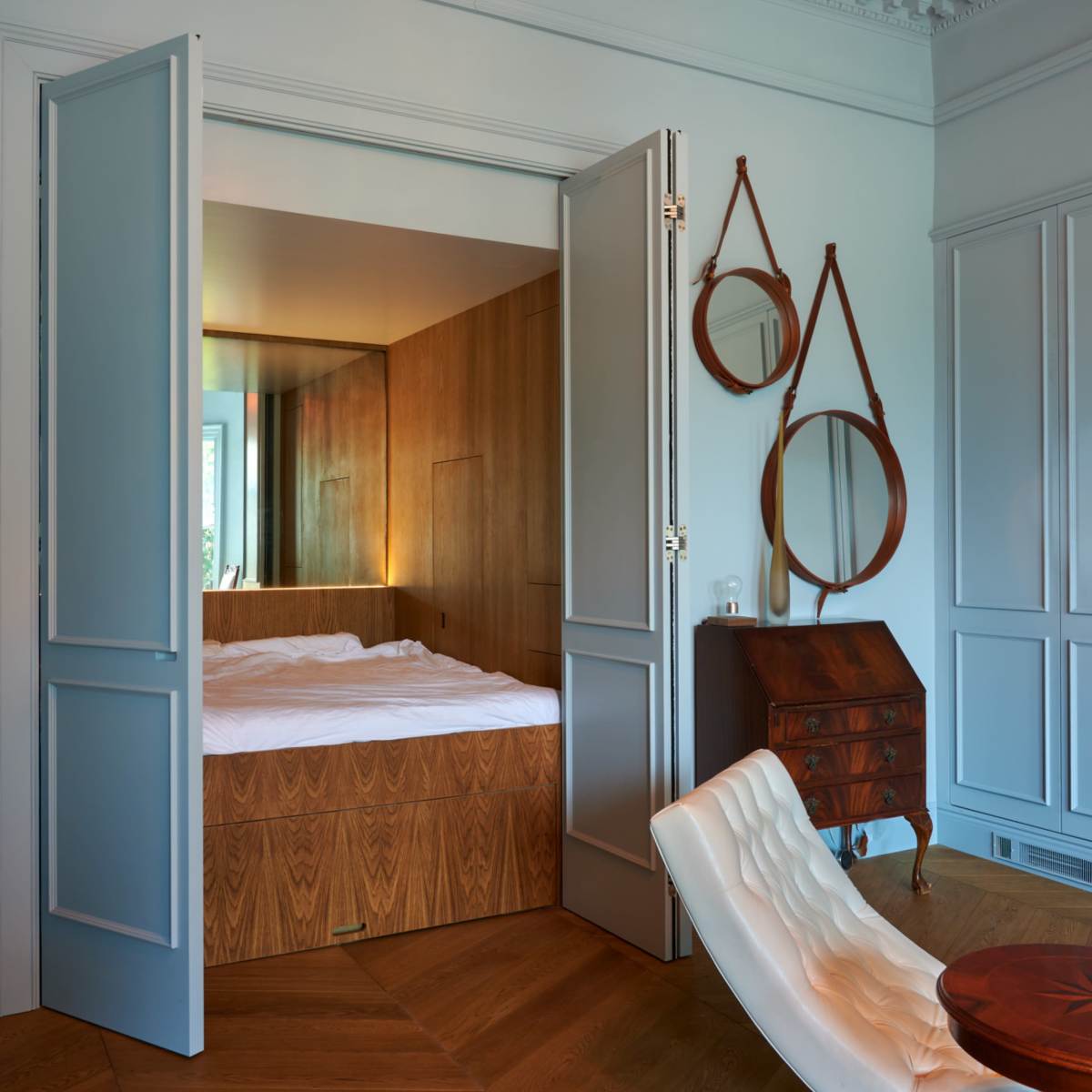 A bedroom is visible through open double doors, featuring a wooden platform bed and contrasting decorative elements in a softly lit modern interior.