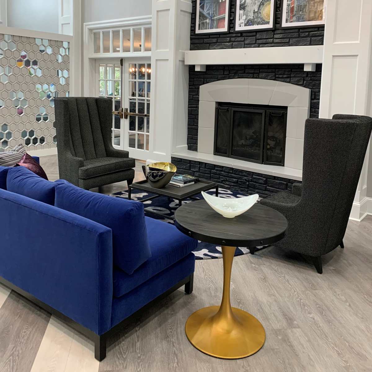 A modern living area with a blue sofa, two gray wingback chairs, a round black table, and a fireplace featuring a decorative wall with framed artworks above it.