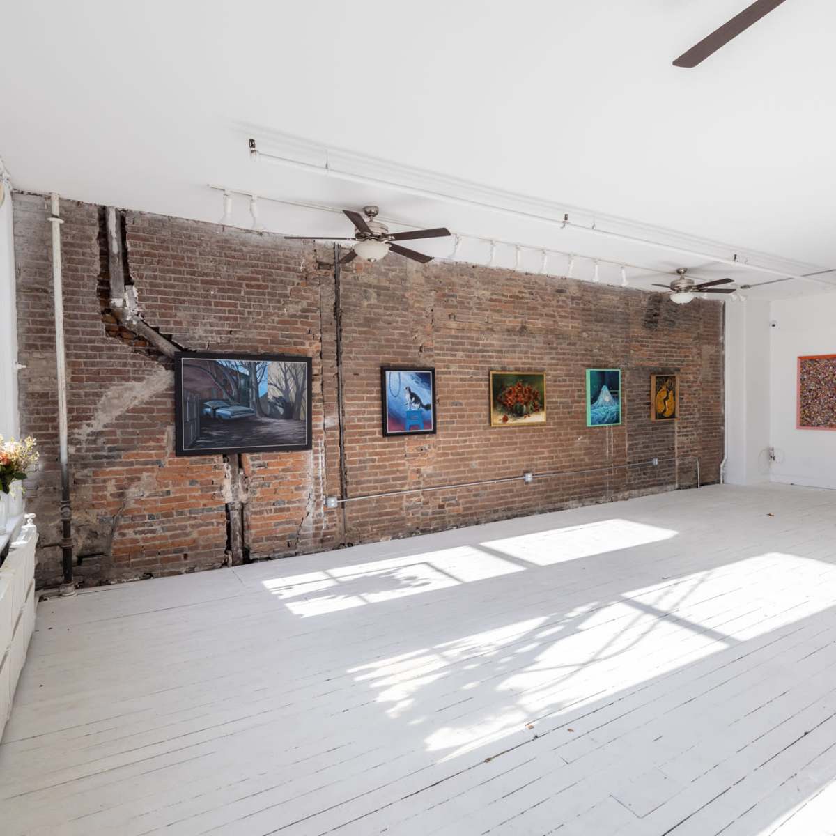 An art gallery with exposed brick walls and white wooden floors, displaying various framed paintings.