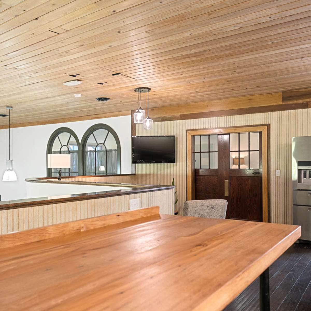 A modern interior space featuring wooden ceilings, a wooden countertop, a set of pendant lights, an arched window, and a kitchen area with stainless steel appliances.