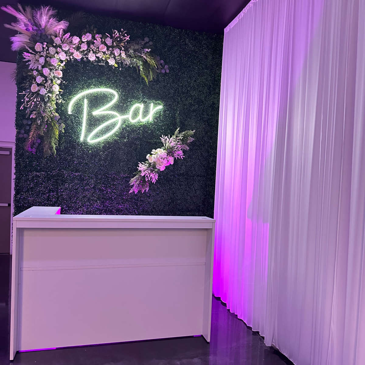 A modern bar area with a white counter, a neon "Bar" sign, and a backdrop of greenery and floral decorations.