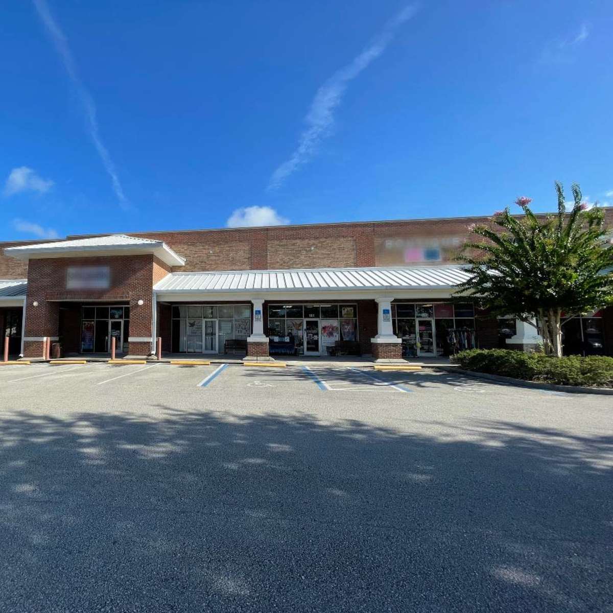 A single-story retail building with a centered entrance and large windows sits in a parking lot under a clear blue sky.