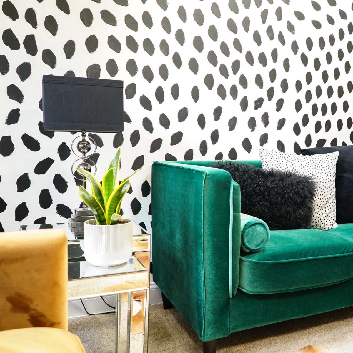 A green velvet sofa with decorative pillows is positioned against a wall featuring a black and white spotted pattern.
