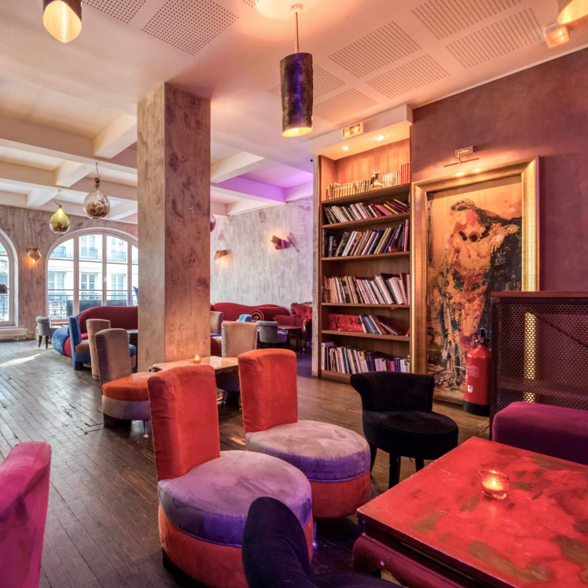 A cozy café interior with colorful armchairs, wooden floors, and arching windows that reveal a view of the outside.
