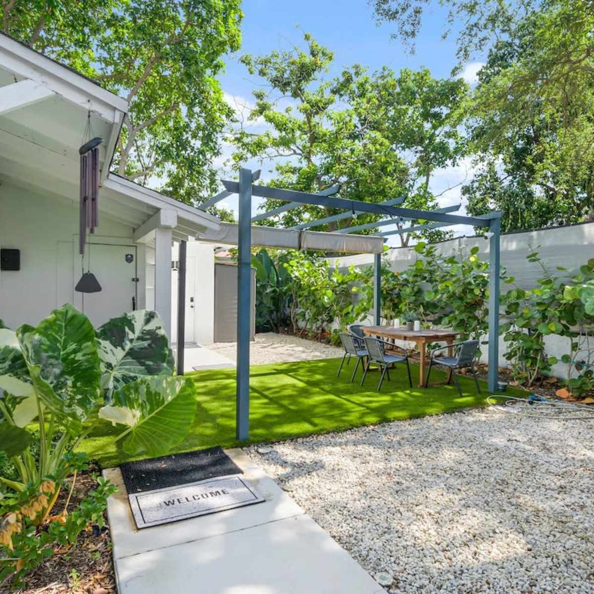A small outdoor seating area with a table under a pergola, surrounded by greenery and gravel pathways next to a house.