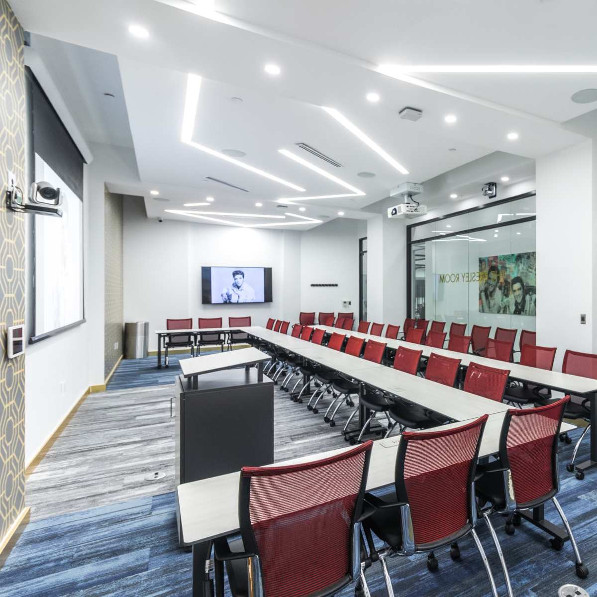 A modern conference room features long tables arranged in a U-shape, with red chairs, large windows, and screens for presentations.