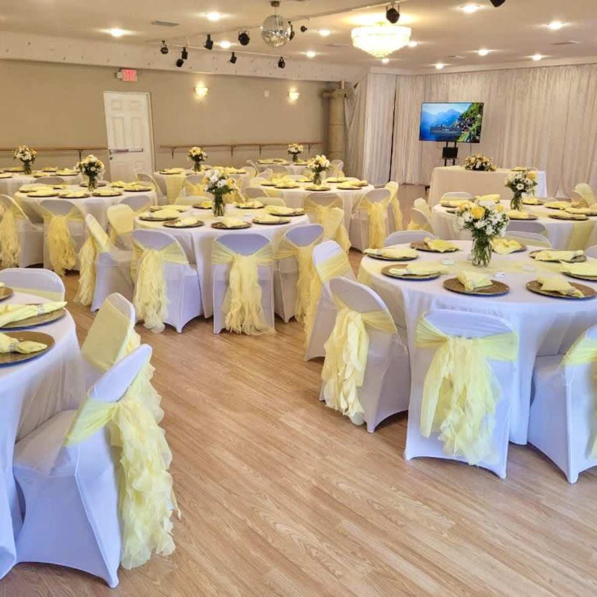 A banquet hall set up for an event with round tables covered in white tablecloths and decorated with yellow-tinted centerpieces, surrounded by chairs draped in yellow fabric.