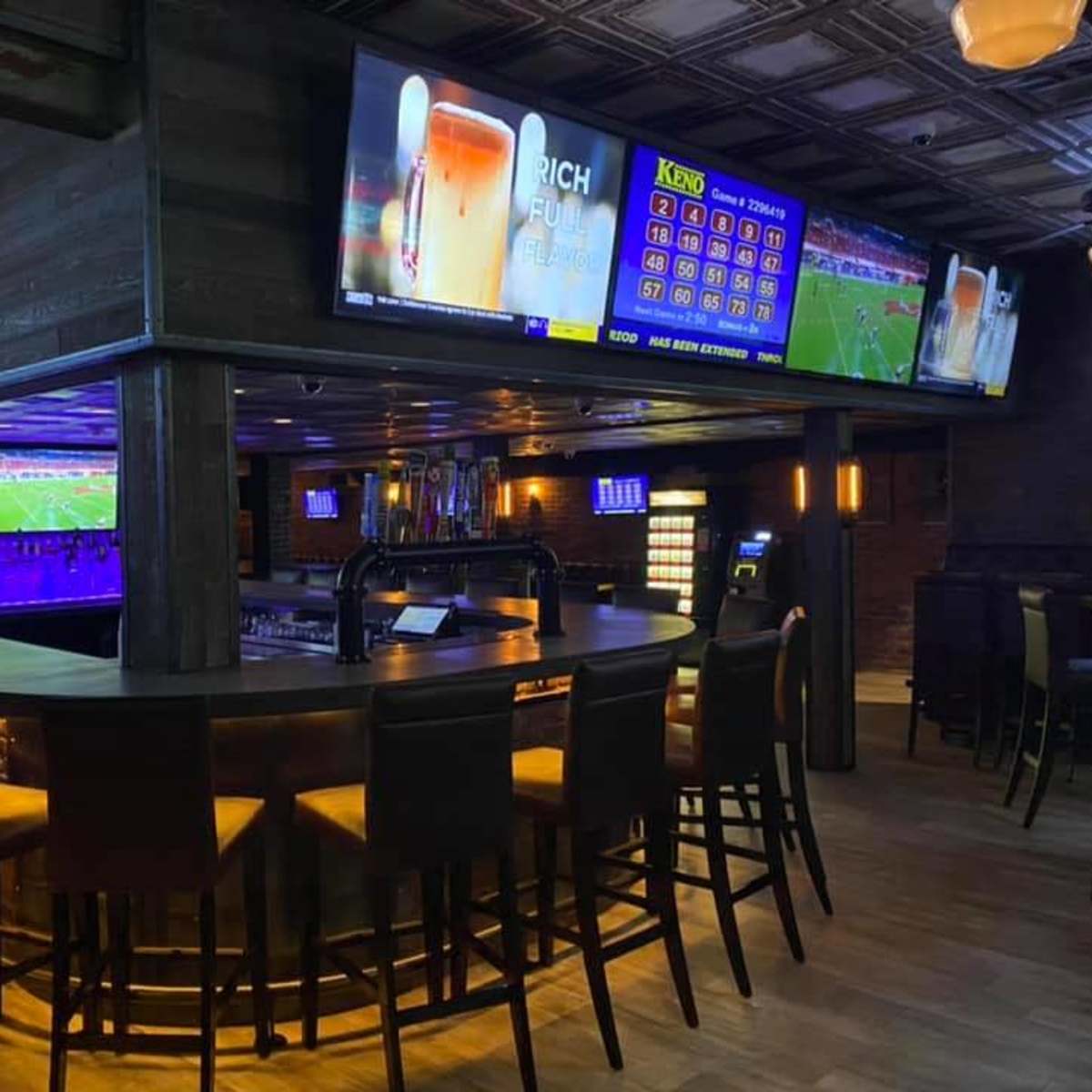 A modern bar interior with a curved counter, high stools, and multiple large screens displaying sports events and games.