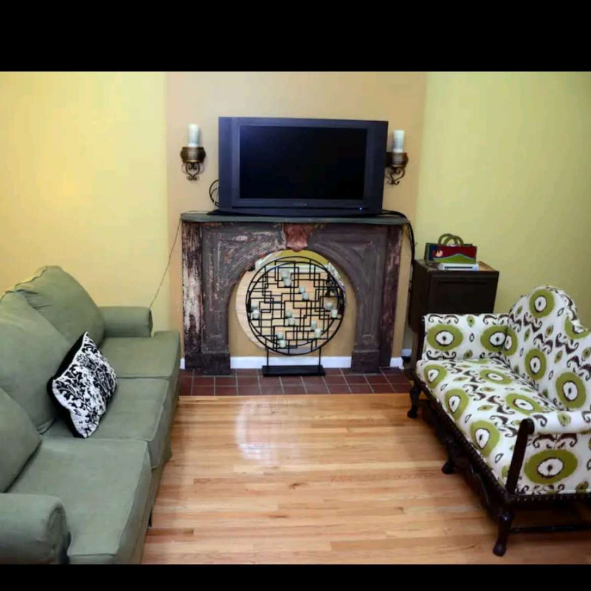 A living room with a green sofa, a patterned armchair, a flat-screen TV mounted above a decorative fireplace, and wooden flooring.