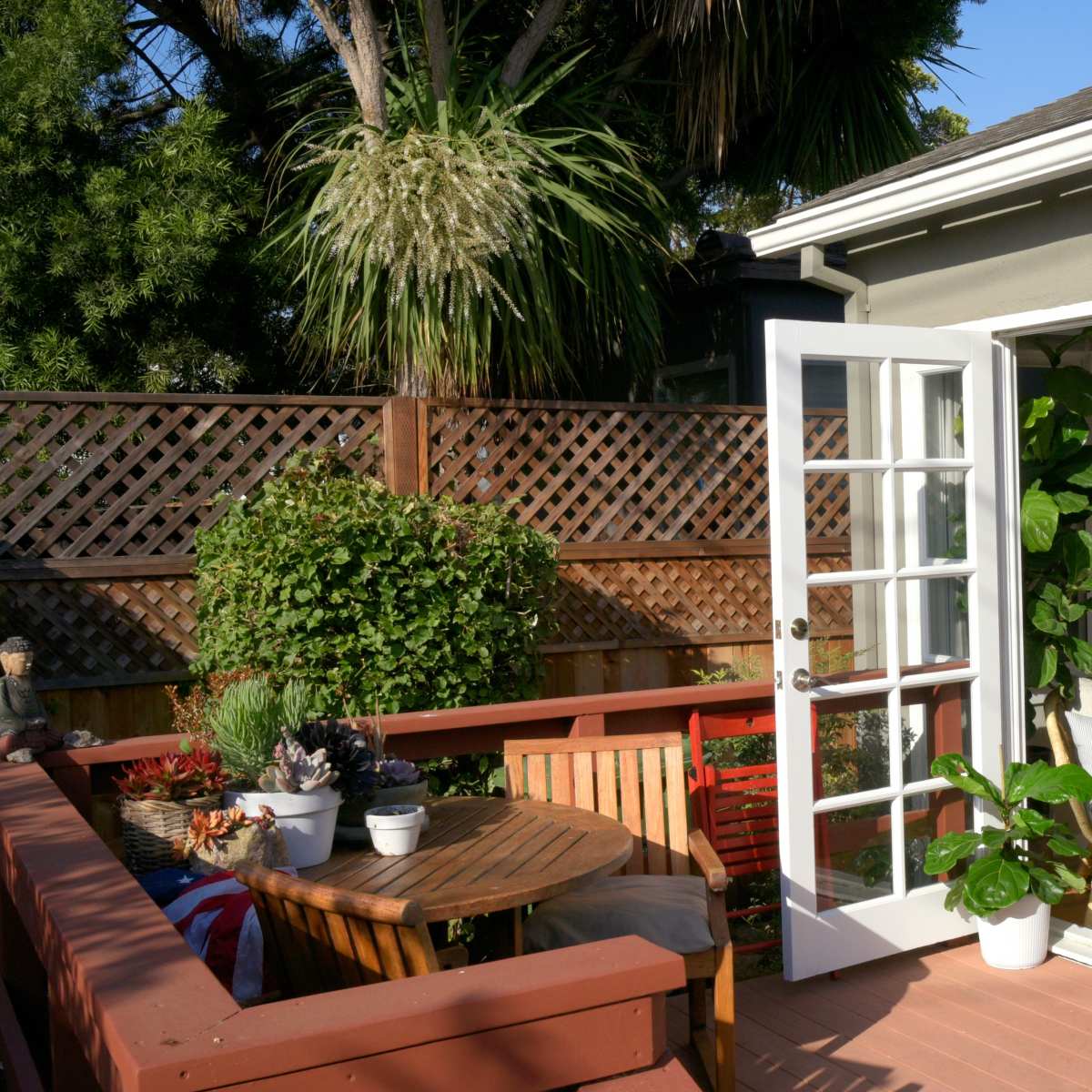 A deck with a round table and chairs is adjacent to a house with open French doors, leading to a cozy indoor space filled with plants.
