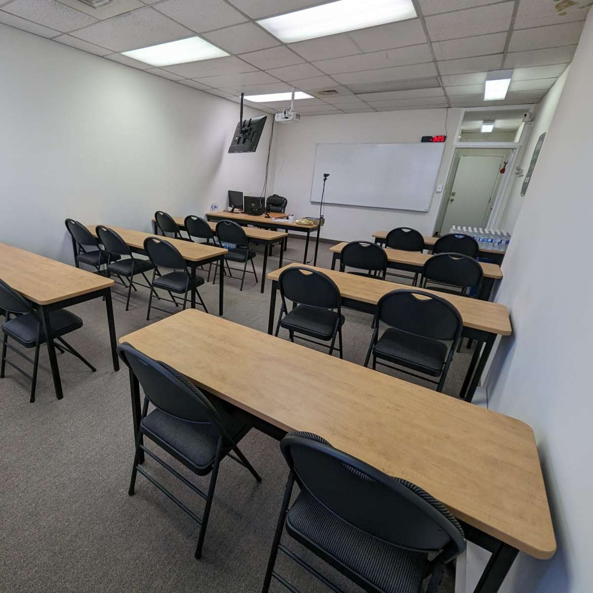 A classroom features several rows of tables and chairs arranged in an organized manner with a whiteboard and electronic equipment at the front.