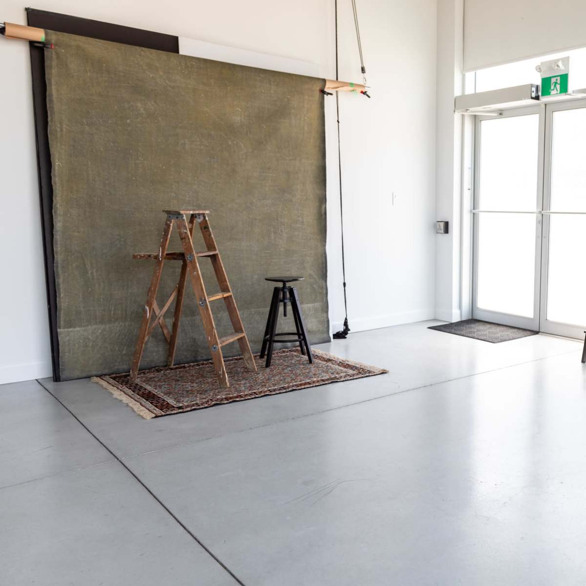 A photography studio featuring a wooden ladder, a stool, a backdrop draped on a frame, and various seating blocks arranged against a wall.