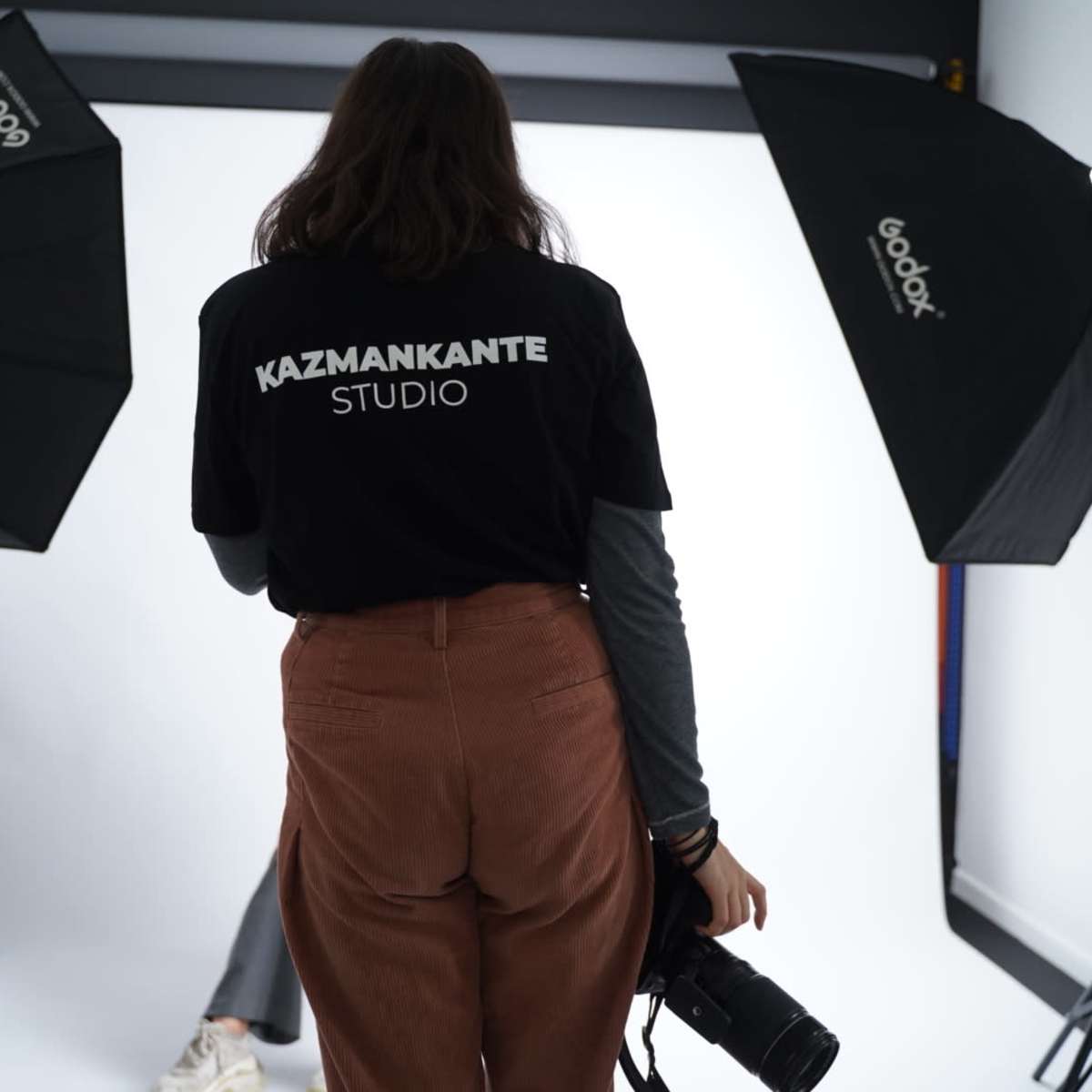 A person stands in a photography studio wearing a shirt that says "KAZMANKANTE STUDIO" while holding a camera, with lighting equipment set up around them.