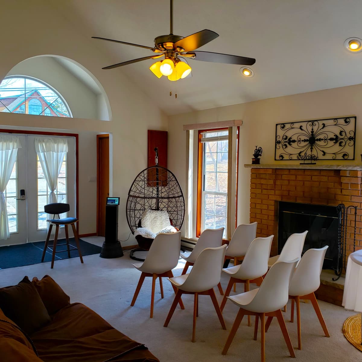 A living room with a circular seating arrangement of white chairs, a fireplace, and sunlight streaming through large windows.