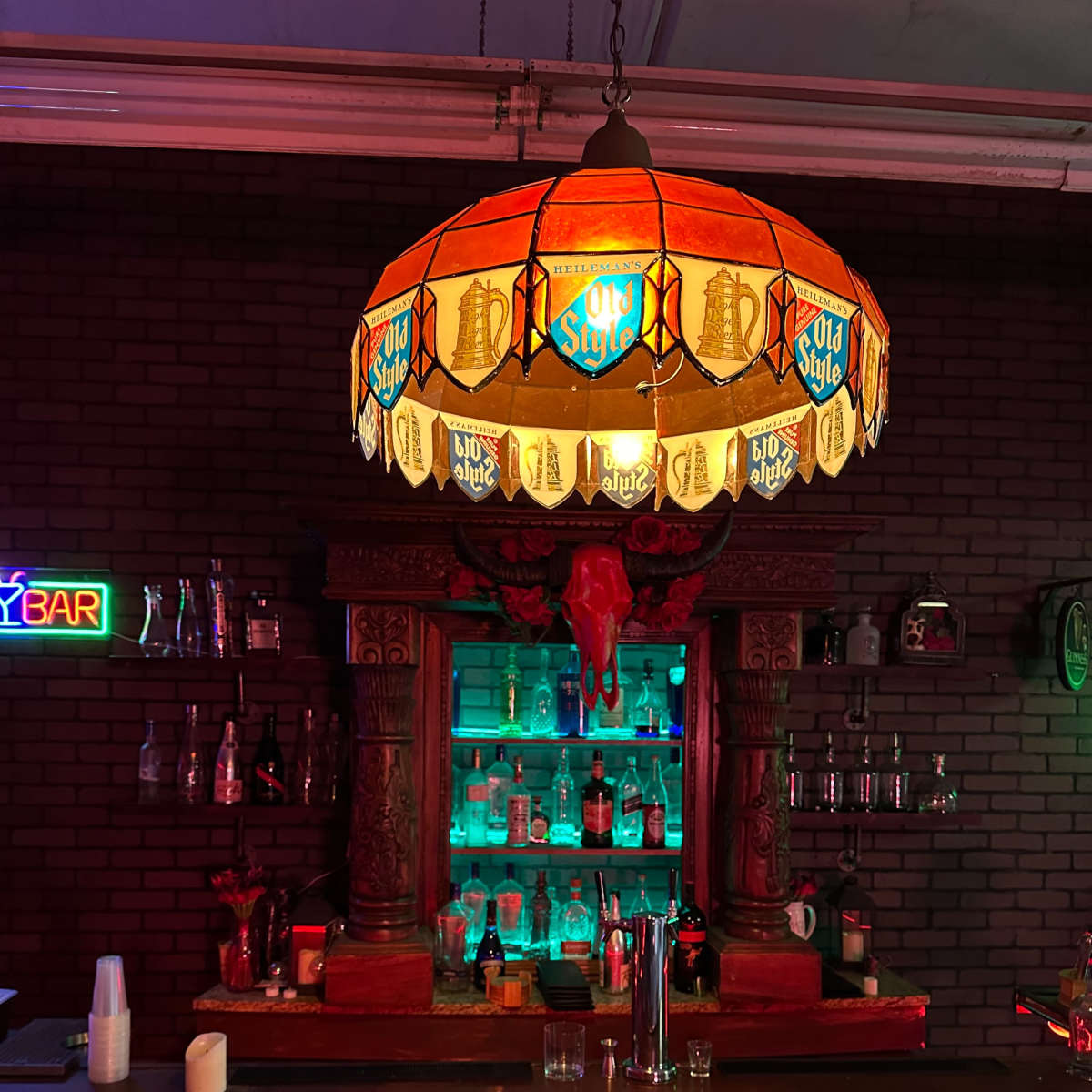 A bar with a wooden counter, colorful lighting, and a prominent stained glass lamp featuring a rustic design above a display of bottles.