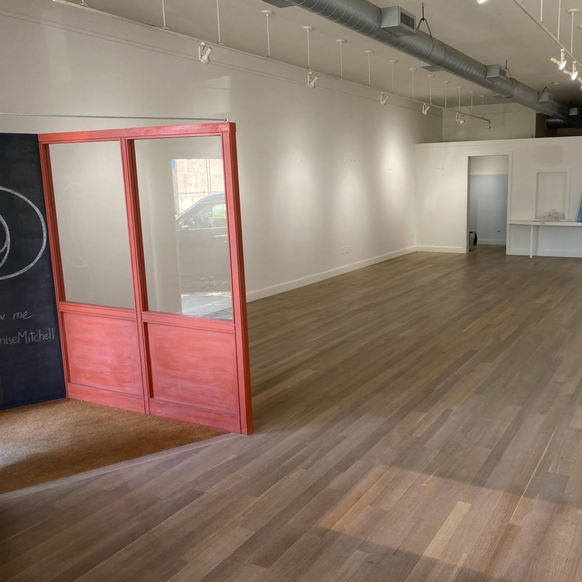 An empty commercial space with wooden floors, white walls, and a glass-paneled door leading to another room.