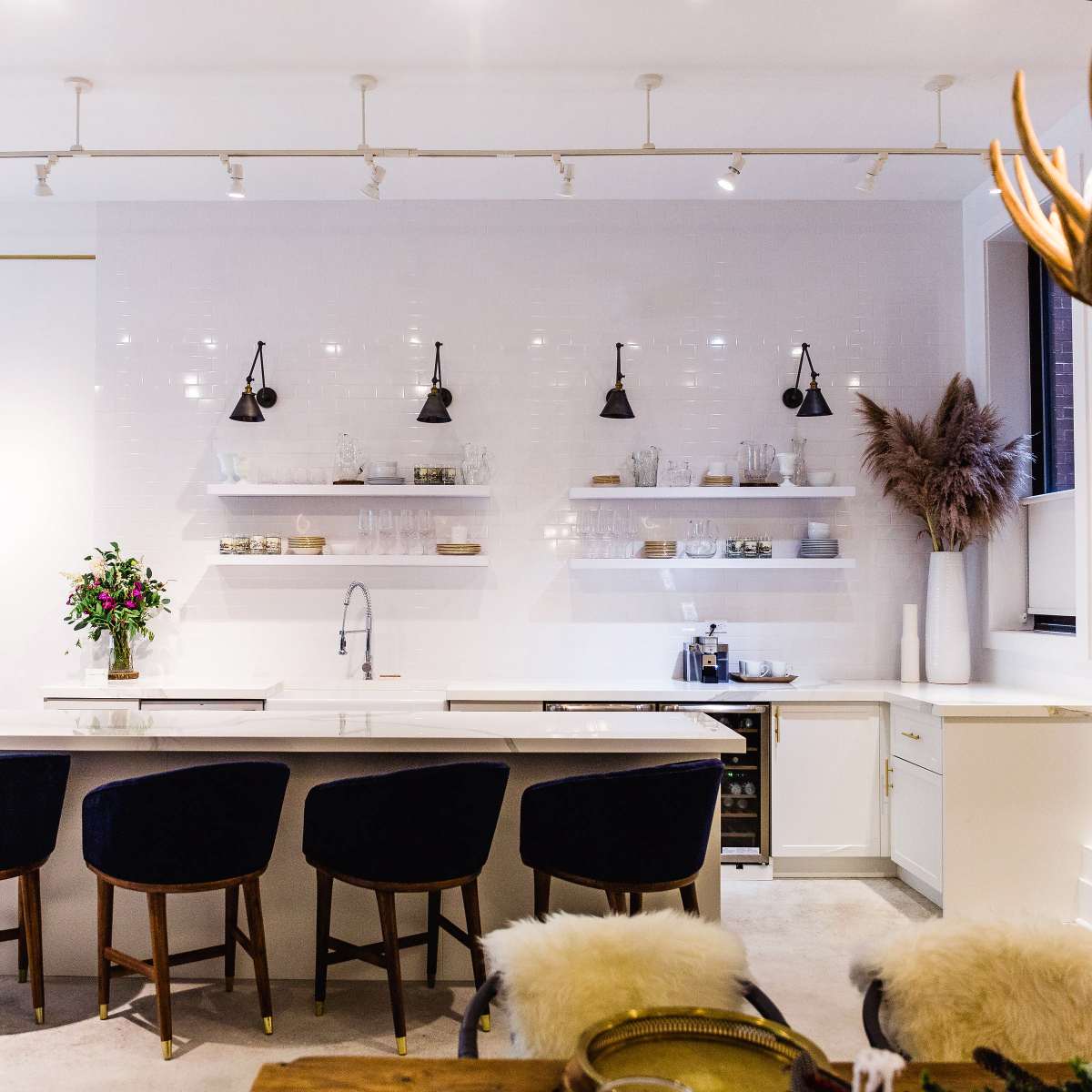 A modern kitchen with a large white island, four dark blue bar stools, open shelving displaying glassware, and a wall adorned with pendant lights and decorative plants.