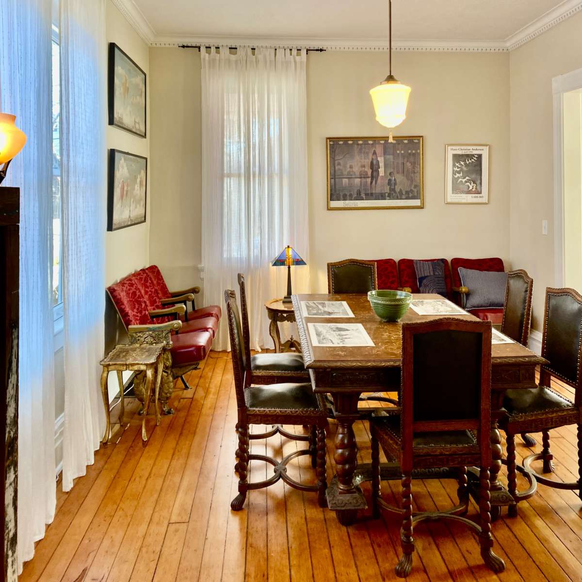 A dining room with a wooden table surrounded by chairs, natural light filtering through sheer curtains, and various artworks hanging on the walls.