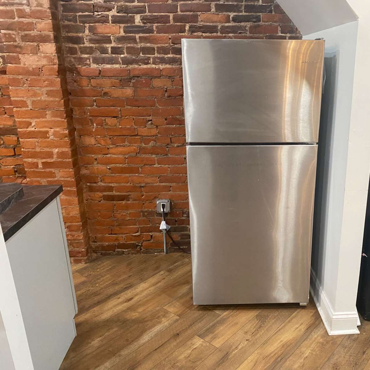 A stainless steel refrigerator is positioned against a brick wall in a corner of a room with hardwood flooring.
