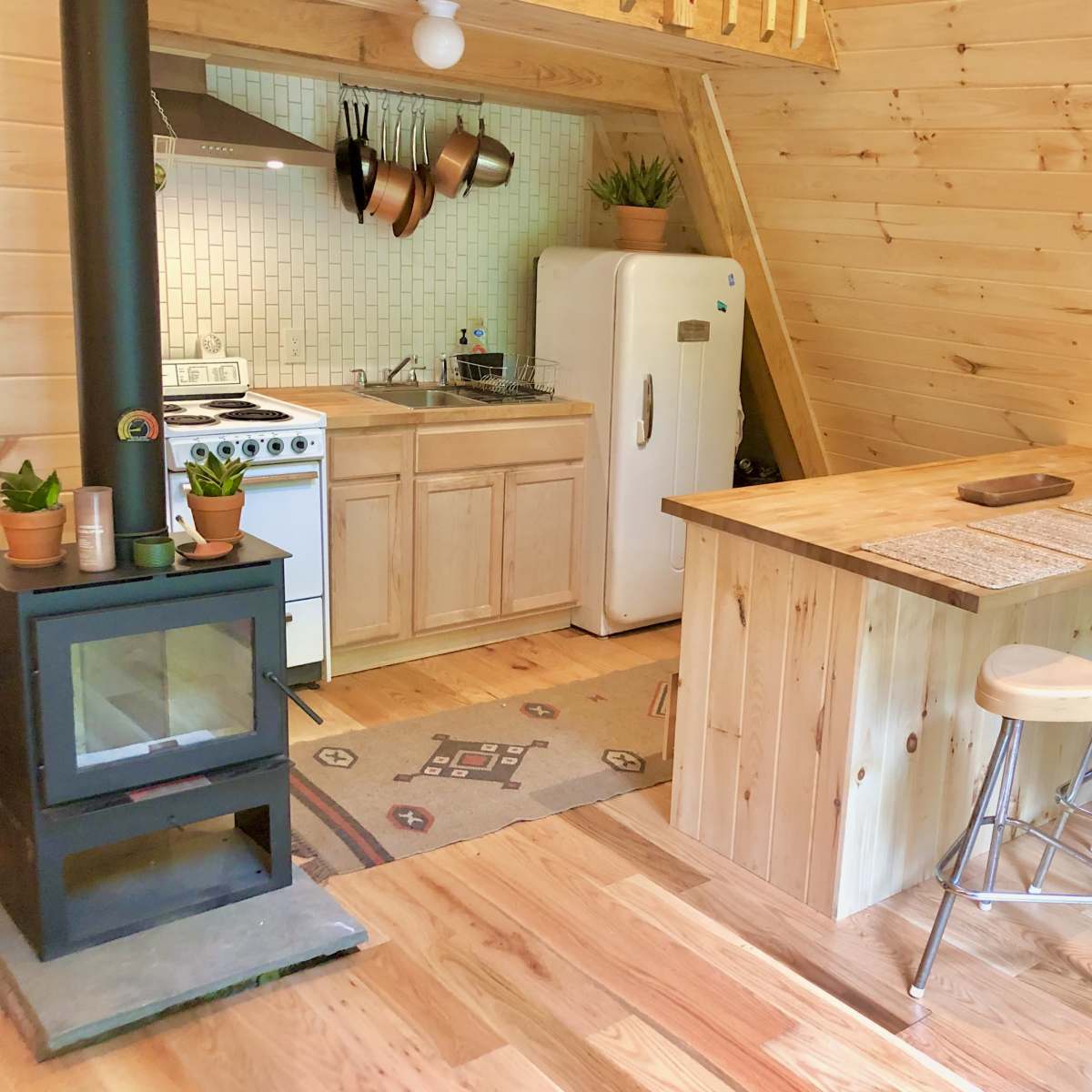 A cozy kitchen area features wooden cabinets, a white refrigerator, and a stove, with a wooden bar and stools beside a small wood-burning stove.