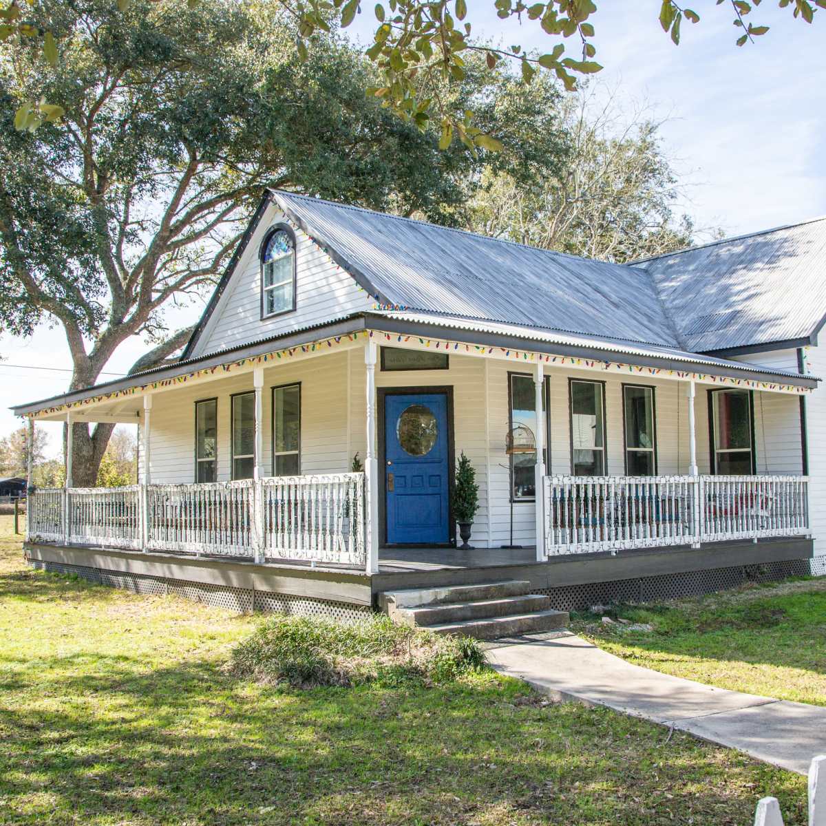 A white, two-story house with a blue door, surrounded by a green lawn and trees.