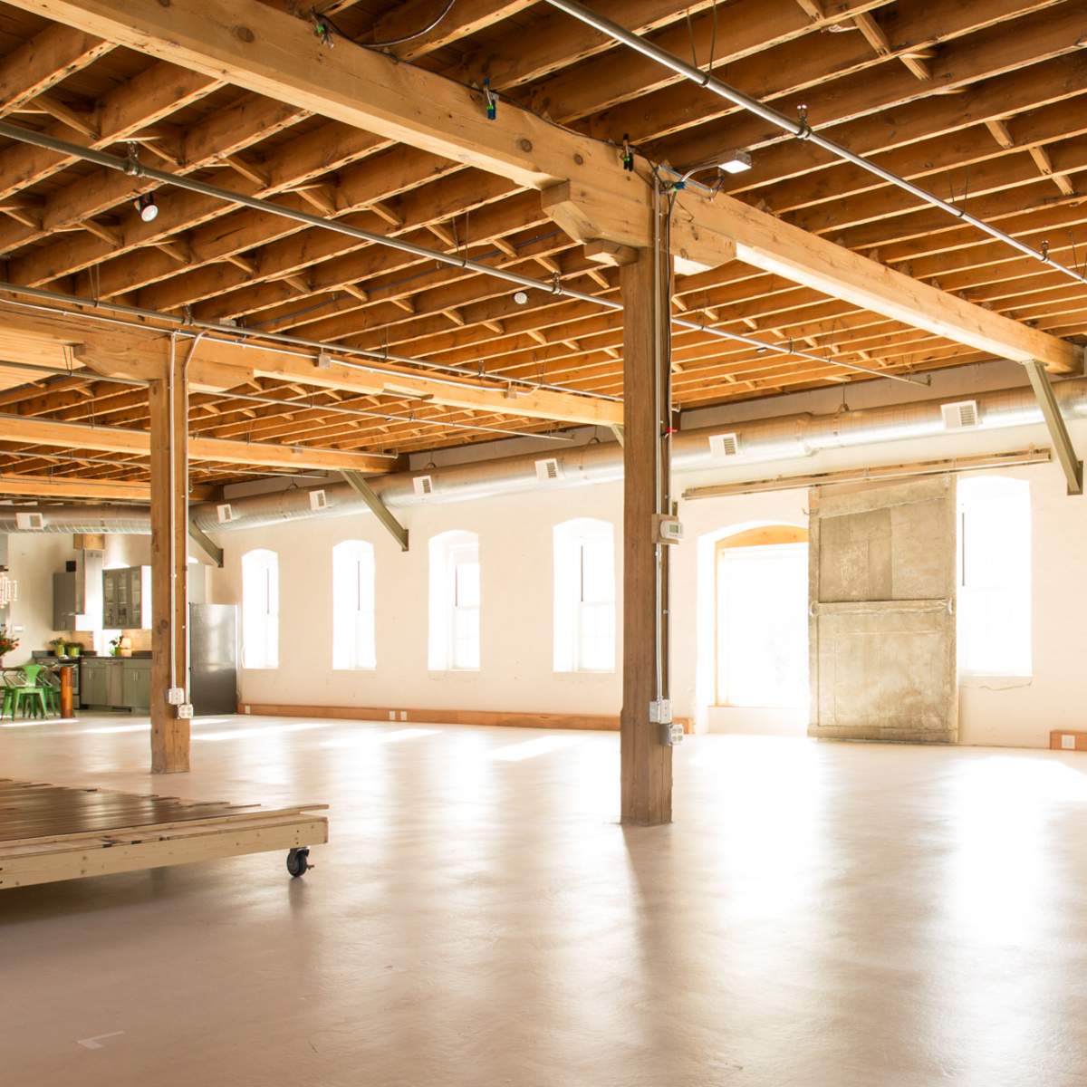 An open, well-lit loft space with wooden beams and a polished floor, featuring large windows and minimal furniture.